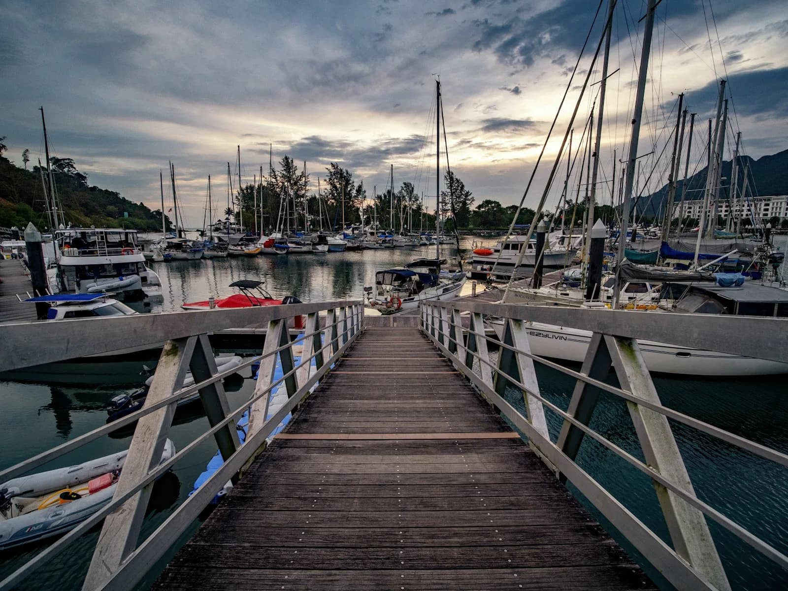 A jetty leading to yachts at a Langkawi marina at sunset, with the island hills behind