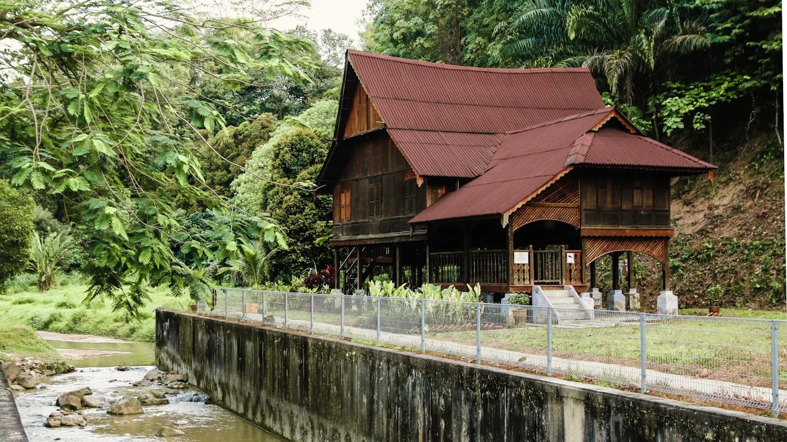 A traditional Malay timber house with a carved verandah beside a forest stream